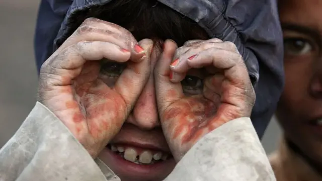 تصویری از یک کودک افغان که انگشت‌هایش را مانند یک عینک دور چشم‌هایش حلقه کرده است. دست‌های او رنگ حنا دارند و لبخند زیبایی بر لبانش نقش بسته.