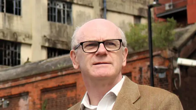 Neil MacDonald aparta la mirada de la cámara hacia la distancia. Es calvo, con algunas canas a los lados de la cabeza. Lleva gafas y una chaqueta de tweed con una camisa blanca debajo. Se ve un edificio antiguo detrás de él.