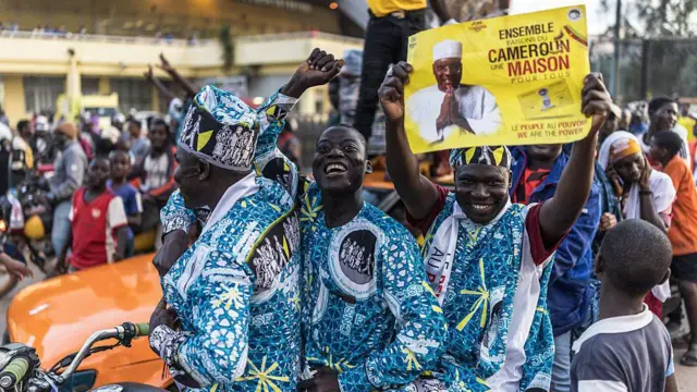 Three men wearing identical blue outfits sit on a motorcycle. One holds up a poster promoting Tchiroma Bakary's candidacy.