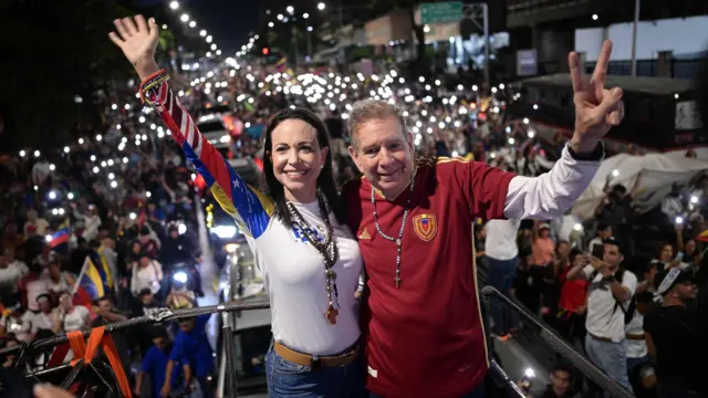 Machado (left) with González standing on a podium on 4 July 2024, with one arm each around one another and their other arm waving. A large crowd is behind them, with many people shining lights. Machado is wearing a white shirt, with the arm red, white and blue. Gonzales is wearing a burgundy shirt with a motif on the left breast.
