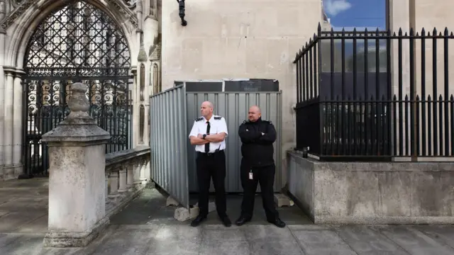 皇家司法院外,兩名身穿制服的保安人員雙臂交叉抱在胸前,站在鋼板前,鋼板遮住了班克斯的壁畫。