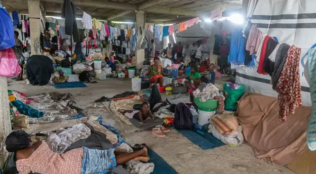 أشخاص في مأوى مؤقت في بورت أو برنس، يجلسون ويستلقون على حصائر على أرضية خرسانية، وفوقهم حبال غسيل معلقة. في الخلفية، تظهر عدة دلاء ماء وأكوام من الأمتعة.