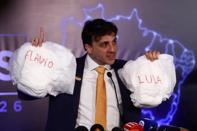 Renan Santos com duas fraldas, uma em cada mão; em uma, está escrito Flávio e, em outra, Lula