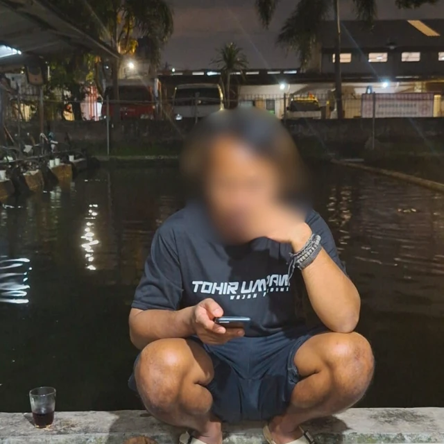 Amir dengan wajah dikaburkan sedang jongkok di pinggir kolam ikan sambil memainkan ponselnya.