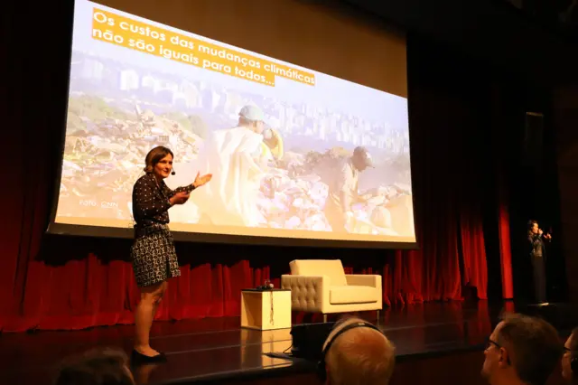 Mulher branca, com roupa preta, fala num palco. atrás uma imagem de homens trabalhando num lixão, com a frase 'custos das mudanças climáticas não são iguais para todos'