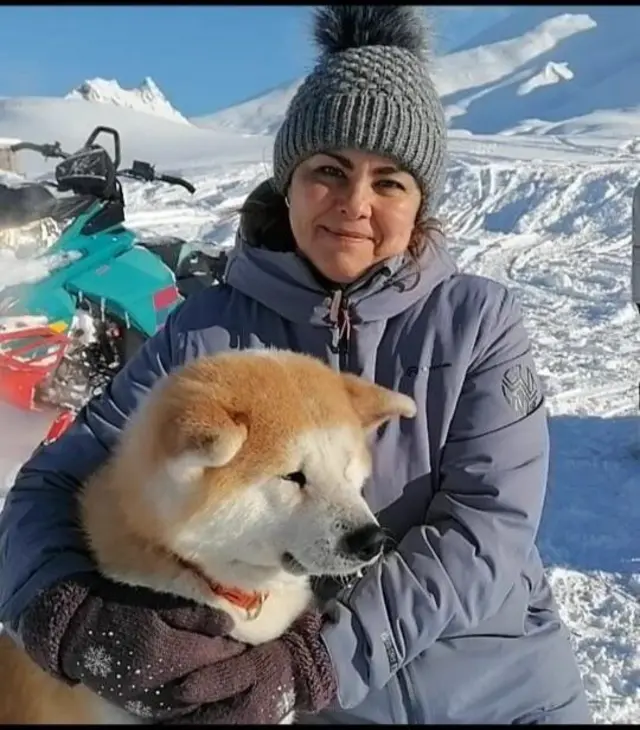 Женщина в теплой одежде улыбается на фоне заснеженных гор и обнимает пушистую собаку