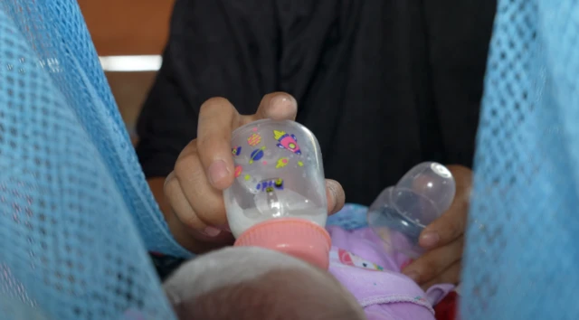 Nurul sedang memberikan susu formula (sufor) dalam botol ke bayi perempuannya, yang ditaruh di timangan gantung