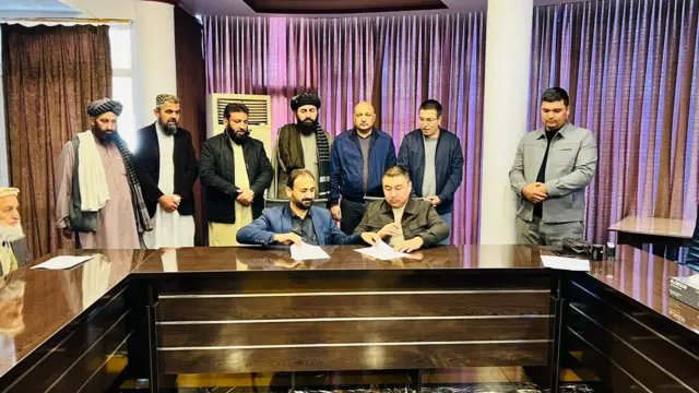 دو مرد در حال امضای قرارداد در پشت میزی نشسته و چندین نفر دیگر در پست آنان ایستاده ‌اند