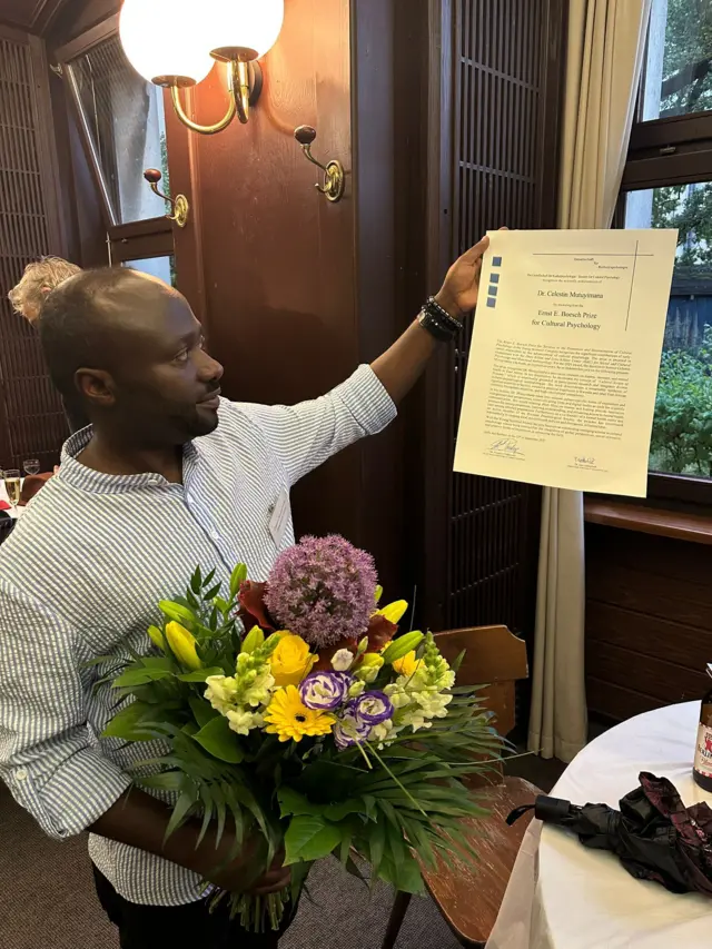 Dr Célestin Mutuyimana afashe inyandiko y'igihembo cye cya 'Ernst Eduard Boesch Prize for Cultural Psychology', afite n'indabo. Ifoto yo muri Nzeri (9) mu mwaka wa 2025, i Berlin mu Budage. 