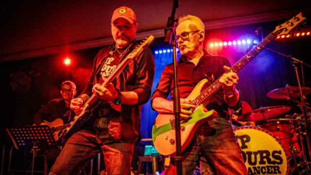 Two men are standing with guitars on a stage, with a microphone in the middle of them. In the background is another man with a guitar, and a pair of drums. 