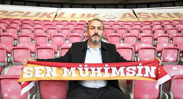 Sebahattin Şirin sarı kırmızılı arka plan üzerine "sen mühimsin" yazılı bir Galatasaray kaşkolunu tutuyor ve tribünde kırmızı koltuklar üzerinde oturuyor