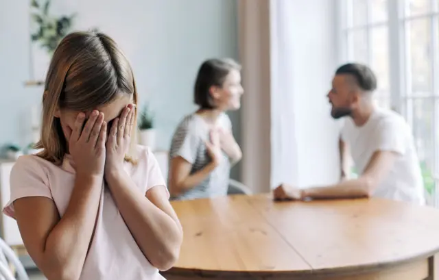 طفلة صغيرة ترتدي قميصاً قصير الأكمام يبدو أنه وردي اللون، وتغطي وجهها بكفيها وتبدو مستاءة من جدال بين والديها اللذين يظهران خلفها جالسين على طاولة خشبية لكن لا تبدو تفاصيل وجهيهما واضحة. الأب والأم شعرهما يميل إلى اللون الكستنائي يرتدي الأب قميصاً قصير الأكمام أبيض والأم قميصاً قصير الأكمام أيضاً ومخططاً باللونين الأسود والأبيض.