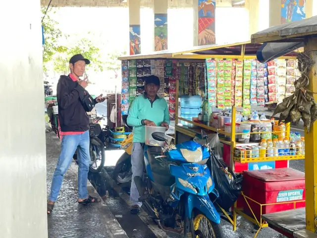 Tafrizal, 58 tahun, menggunakan topi dan kaos kerah hijau sedang mengambil sesuatu dari dalam wadah plastik. Ia berada di samping gerobaknya yang dimodifikasi dengan sepeda motor dipenuhi dengan rencengan minuman instan.