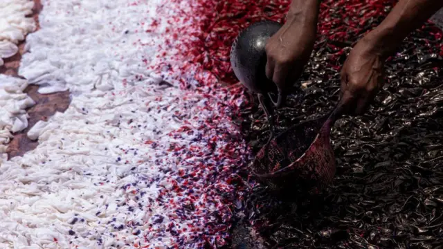 Hands pour a dark liquid from a small bowl onto a large piece of fabric spread across the frame, creating a dense pattern of red, purple and black dye. The fabric fills the foreground and background, with sections of white cloth on the left gradually blending into darker dyed areas on the right.