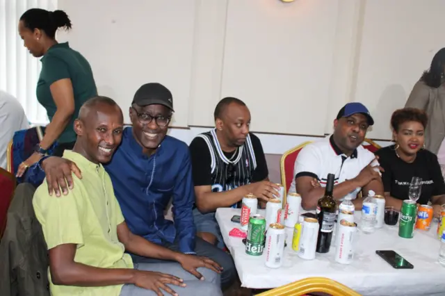 Abantu baryohewe bicaye, imbere yabo hari amacupa, iruhande hari umuntu arimo kugenda