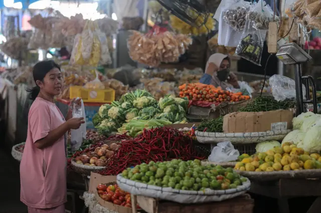 Seorang perempuan berkaos merah dengan rambut dikuncir sedang berada di depan barang dagangan berupa cabai, kubis, wortel, tomat dan jeruk di sebuah pasar. 