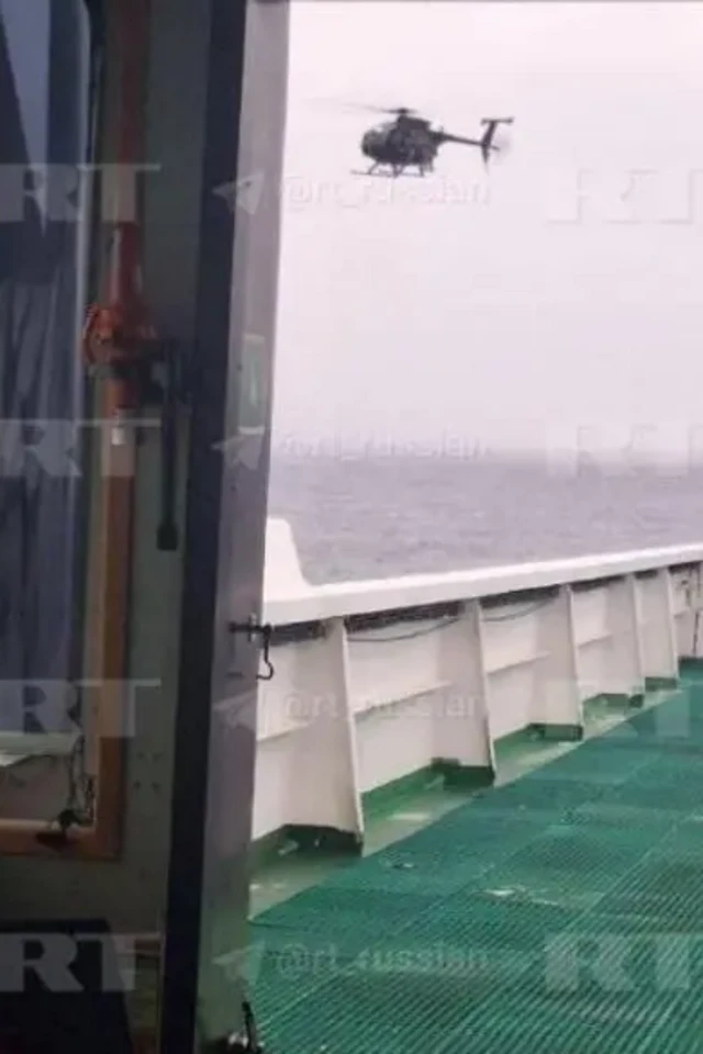 Вигляд зсередини судна на зелену металеву палубу; за бортом у небі вертоліт зависає над водою.