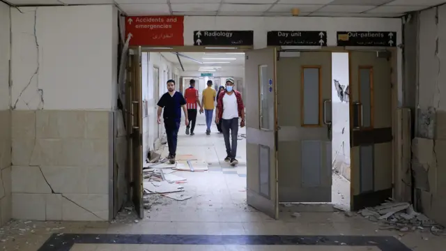 Image of the corridor of a hospital, showing cracked walls, rubble and people walking