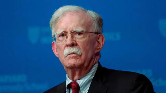 Close-up image of John Bolton, e dey look straight ahead. E wear glasses, black blazer, stripped blue and white shirt and a red tie. 