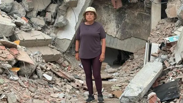 María Lourdes Pérez, vestida con un pantalón y camisa marrones y un gorro beige. Detrás de ella hay una montaña de escombros, piedras, partes de una edificación, ladrillos.