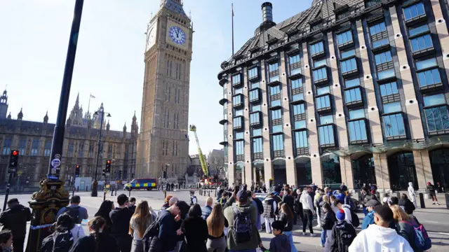 A cherry picker is parked beside the clock tower at Parliament, as a crowd of people stand some distance away. Portcullis House, a parliamentary office building, is on the right of the picture.