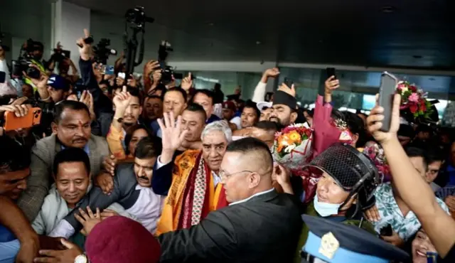 देउवा विमानस्थलमा ओर्लिएपछि हात उठाएर समर्थकहरूलाई अभिभावन गर्दै। क्यामरा बोकेका मानिसहरुको भिड छ।