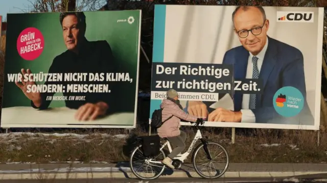 Mulher de bicicleta passando em frente a outdoor com fotos de Friedrich Merz e Robert Habeck