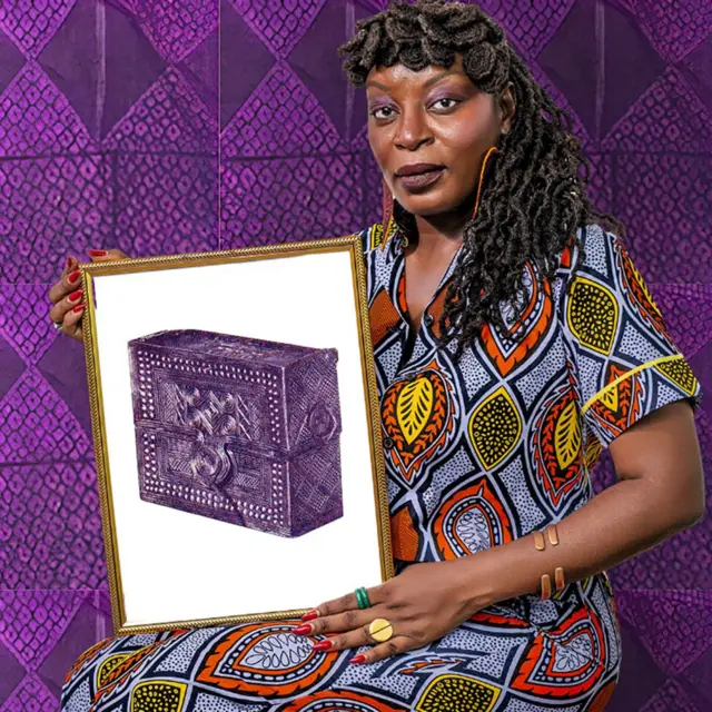 Samba Yonga, du Women's History Museum of Zambia, brandit un cadre montrant une photo d'une boîte à outils de chasseur en bois portant une inscription ancienne. Elle porte de longues tresses, un fard à paupières rose, du vernis rouge sur les ongles et une robe à imprimé africain jaune, orange, noir et bleu. Elle est photographiée sur un fond d'imprimé africain violet et noir.