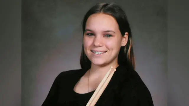 Taylor, 14 yaşındayken resmi bir portre için yumuşak gri bir stüdyo fonunun önünde duruyor. Sade siyah kısa kollu bir bluz giymiş ve göğsünün üzerinde dik duran iki tahta davul tokmağı tutuyor. Uzun koyu saçları omuzlarından aşağı dökülüyor ve dişlerindeki metal teller görünürken gülümsüyor.