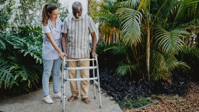 Homem negro idoso caminha com andador, ao ladocomo apostar no brasileiraouma mulhercomo apostar no brasileiraouniforme