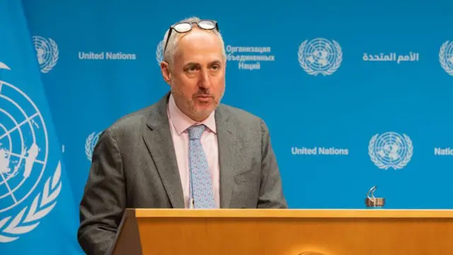 Stephane Dujarric speaking at a new conference. He is standing behind a lectern marked with the UN seal and is wearing a grey suit. 