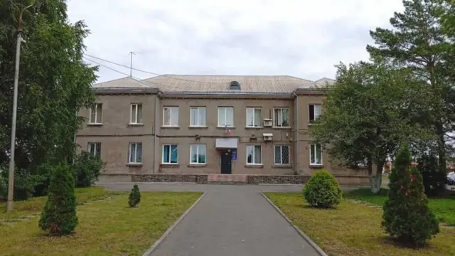 На заднем плане серое двухэтажное здание, на переднем плане — асфальтовая дорога, ведущая к зданию, газон, кусты и деревья по бокам.