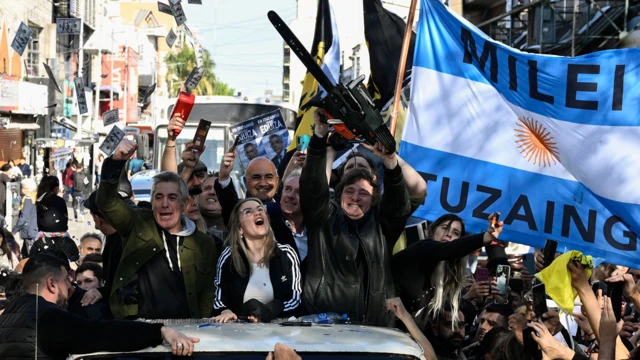 Javier Milei levantando uma motosserra enquanto está em cima de um carro desfilando pela rua. Há vários apoiadores em volta