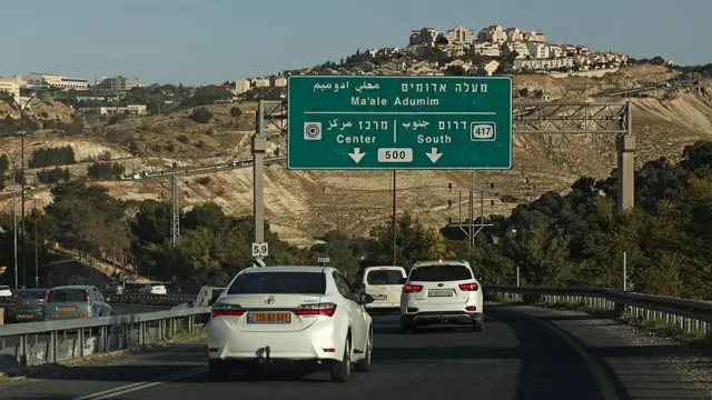 Varios vehículos circulan por una carretera y pasan bajo un cartel que indica el camino al asentamiento de Maale Adumim, que puede verse al fondo de la imagen, sobre una colina.