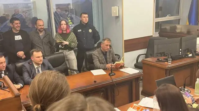 За столом на задньому плані сидять три прокурори. Крайній лівий у смугастому бежевому піджаку Олександр Ганілов