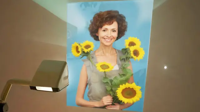 Una imagen de IA de una mujer con cabello corto y rizado, sosteniendo girasoles, proyectada sobre una pared, ella sonríe y mira directamente a la cámara.