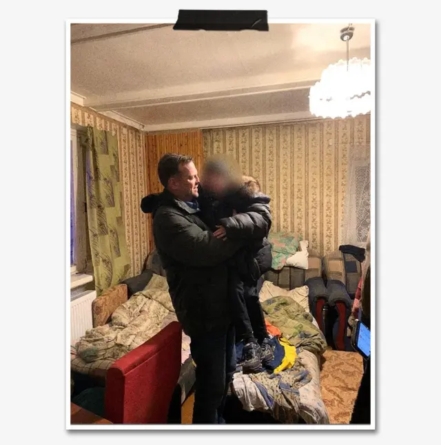 Иван Семенихин — улыбающийся мужчина в темной куртке — стоит в комнате и держит на руках ребенка, его лицо размыто. На заднем плане кровать с одеялом, люстра под потолком.
