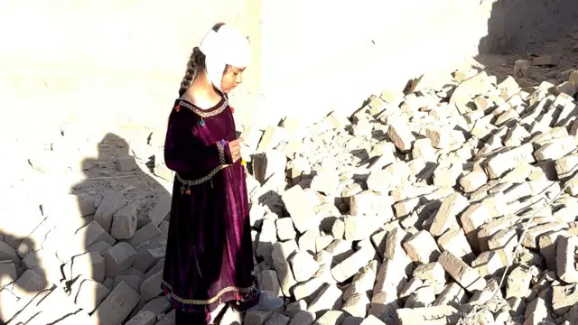 دختر ۶ ساله که در حملات هوایی پاکستان سرش زخمی شده و با بانداژ بسته است، بالای خشتهای فروریختهای خانهاش ایستاده و به فکر فرو رفته است. ۱۳ مارچ ۲۰۲۶