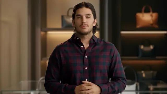 An AI-generated image that shows a man standing inside a handbag shop, wearing a long-sleeved checked shirt. His hands are clasped in front of him and he appears to be speaking. His shoulder-length dark hair is brushed back over his head and he has dark stubble on his face. Behind him are handbags arranged on shelves. They each have their own shelf and are individually lit.