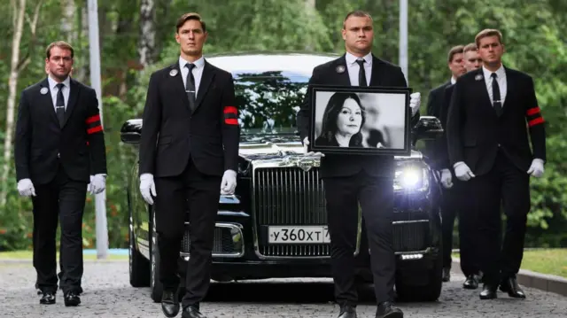 Четыре мужчины в траурных костюмах идут перед катафалком, один несет портрет женщины 