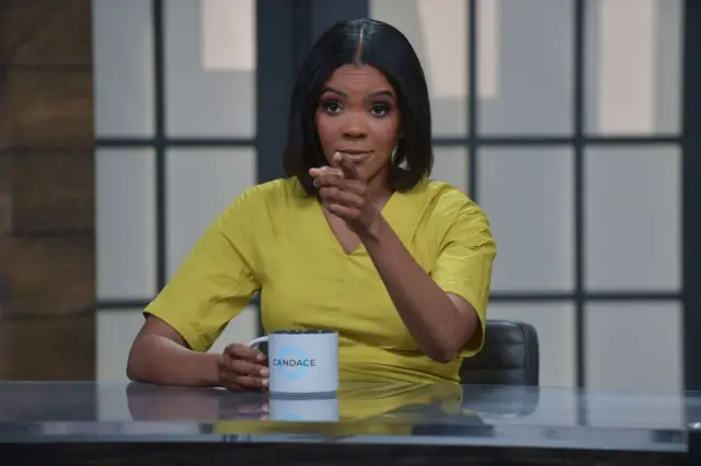 Owens atrás de uma mesa, apontando para a câmera, segurando uma caneca com a inscrição 'Candace'