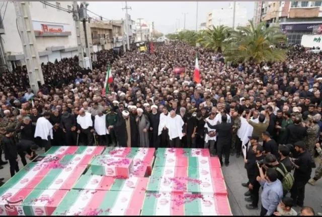 تعدادی تابوت با پرچم‌های جمهوری اسلامی و مردمی که در اطراف آنها برای سوگواری و تشییع گرد آمده‌اند