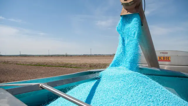 Синие гранулы высыпаются из железной трубы в кузов серебристой машины на фоне свежераспаханного поля под блеклым полуденным небом