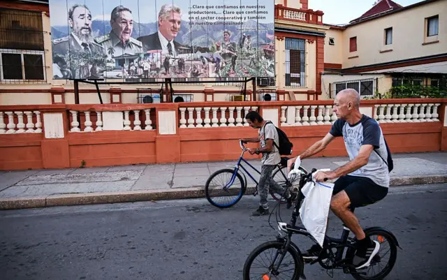 Un hombre en bicicleta pasa delante de un mural con los rostros de Miguel Díaz-Canel, Fidel y Raúl Castro.
