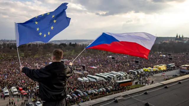 Чоловік на даху будівлі над величезним натовпом протестувальників і тримає два великі прапори – Чехії та ЄС. Внизу видно щільно заповнене людьми поле, припарковані автобуси та техніку. На горизонті – міський краєвид і похмуре небо.