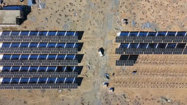 A birds-eye view of five rows of solar panels on the left above the light brown-coloured ground. On the right, two rows of solar panels are complete, with poles visible along three more rows where panels are yet to be installed. 