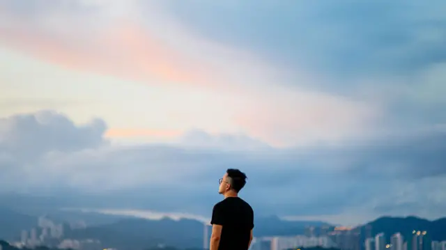 Homem jovem branco de costas, de cabelo curto, olha para um céu cheio de nuvens