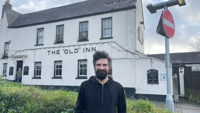 Ryan Bray frente a su pub, el "Old" Inn. Es un hombre barbudo que lleva una sudadera negra con capucha.