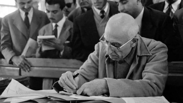 İran Başbakanı Muhammed Mosadeq, arkasında takım elbiseli adamlar varken bir masada oturmuş belgelere bakıyor.