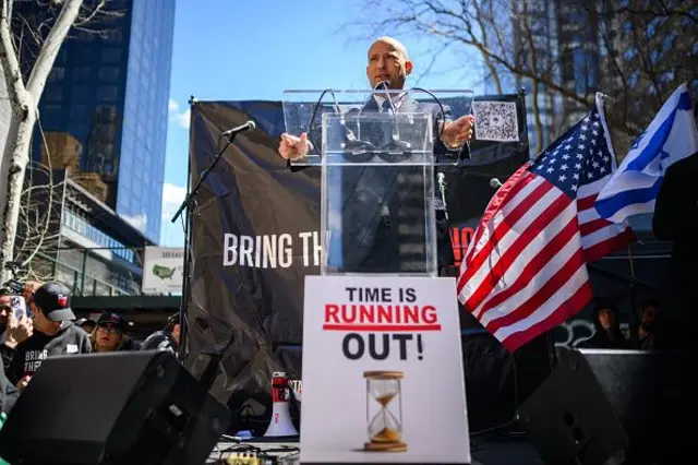 Bennett Zaman Tükeniyor yazılı bir kürsüde konuşma yapıyor yanında ABD ve İsrail bayrakları var. 